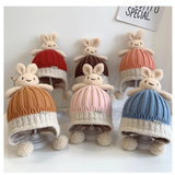 Six Pret my baby “Thick warm woollen rabbit top beanie cap” hats in red, brown, yellow, pink, blue, and orange are displayed. Each woollen beanie has a plush rabbit with floppy ears and dangling limbs. A sign reads “SOFTLY SPECIAL.”.