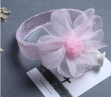 The Pret my baby Lace headband with large organza flower features a delicate pink lace band and flower detail, ideal for elegant outfits. Displayed on a gray surface with a partially visible black-and-white photo card underneath.