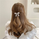 A woman with wavy light brown hair styled half-up, secured with the Pretmebabe Evening metal bow buckle with asymmetric pearls, wears a white ruffled blouse and is seen from behind in a softly lit, minimalist room.
