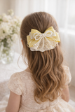 A young girl in a cream lace dress wears Pret my baby's Multiple bow with doiley lace fascinator hairclip, featuring yellow and white bows, pearls, and secured by an alligator pin, as she sits near a vase of white flowers.