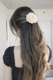 A woman with long, wavy brown hair styled half-up wears a Pret my baby Large crochet rose bloom hairpin in cream. She is dressed in a black short-sleeved top and faces away from the camera in a bright room.