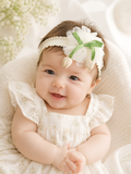A smiling baby in a floral dress wears the Pret my baby Fancy Fascinator Little Tulips Baby Headband, featuring green ribbon and flower decorations, while sitting on a soft cream-colored blanket with a blurred floral background.