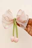 A hand holds the Pret my baby Medium fascinator tulip bow—a sheer, light pink organza bow with green ribbon tails tipped with pink, cherry-like buds and delicate tulip-inspired pink-edged trim.