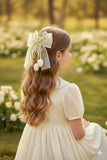 A young girl with wavy brown hair sits on the grass, wearing a cream-colored dress and a Pret my baby Tulip with pearls large bow fascinator hairclip. Blooming white flowers surround her in the background.