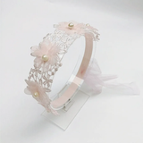 The Pret my baby Lace headband with soft flowers and pearls features light pink floral details and pearl accents, displayed on a clear stand against a white background—ideal for elegant outfits.