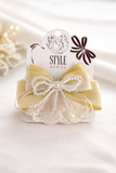 The Pret my baby Multiple bow with doiley lace fascinator hairclip features yellow and white bows, pearl accents, and delicate doiley lace, displayed on a branded card against a soft white textured background.