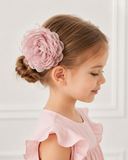 A young girl with light brown hair in a low bun wears the Pret my baby Fascinator extra large flower hairclip and a light pink ruffled dress, smiling gently while facing sideways against a light background.
