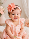 A happy baby sits indoors, wearing a pink dress and the Pret my baby Soft cotton everyday baby headband with a large crush flower, surrounded by soft pastel floral decorations.