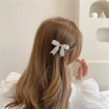 A woman with long, light brown hair wears the Pretmebabe Evening metal bow buckle with asymmetric pearls, pairing it with a white puffy-sleeved top as she sits in a softly lit, minimalistic room, her face turned away from the camera.