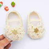 A pair of cream Rosette bellies with star from Pret my baby, featuring gold glitter stars and bows with central gold stars. One shoe is held by someone, with small plants in the background—an ideal charming baby gift.
