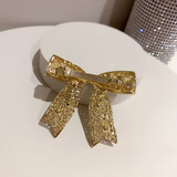 The Pretmebabe Evening Metal Bow Buckle with Asymmetric Pearls, a gold textured hair clip, sits on a white rounded surface as a stunning statement piece. A sparkling rhinestone-covered cylinder in the background adds extra glamour.