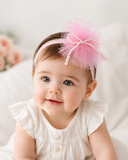 A baby smiles gently while wearing Pret my baby's Fancy fascinator pearls on tulle baby headband and a white dress, sitting on a soft surface.