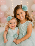 Two young girls in matching mint green dresses and the pretmybaby Flower Bloom Fascinator Sibling Set in sea green smile at the camera. The older girl hugs the baby while pink and white balloons and fairy lights fill the festive background.