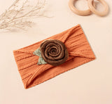 The Pret my baby Soft cotton baby headband with rose - brown, featuring a textured orange knit, large brown fabric rose, and green leaves, is displayed on a beige surface with wooden rings and dried plant accents.