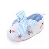 The "Embroidery bellies with stripe bow - white" by Pret my baby are light blue baby shoes with pink floral embroidery, a large blue-and-white striped bow, and a non-slip sole, shown on a white background.