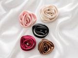The Pret my baby Satin Rose Fascinator Clip features five rose-shaped hair clips in pink, cream, black, red, and brown with pearl accents—perfect for vintage charm or as an adorable baby girl accessory. Displayed on white silky fabric.