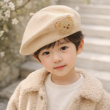A young child with short dark hair wears the Pret my baby Soft Woollen Knitted Beret Cap with a teddy bear design, paired with a matching fuzzy jacket, standing outdoors near stone steps and flowering branches.