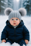 A baby in a navy snowsuit and a Pret my baby Thick woollen double pompon beanie cap with two large furry poms sits on snowy ground in the forest, gazing at the camera with blue eyes and rosy cheeks.