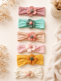The Pret my baby Pack of 6 everyday flower headbands features six colorful, textured flower bands arranged vertically on a light surface with dried flowers for an elegant display.