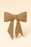 The Pretmebabe Midnight Sparkle Luxe Bow is a gold, rhinestone-encrusted brooch that makes a stunning statement accessory for evening outfits, showcased on a soft beige background.