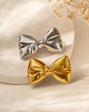 The Pret my baby Pack of 2 metallic bow hairclips, featuring silver and gold bows, is showcased on a white marble surface with dried flowers, adding an elegant shimmer to your hairstyle.