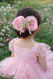 A young girl with brown hair sits in a flower field, her back to the camera, wearing a pink dress and the Pret my baby Tulle curve bow with tulip and pearl ingrain fascinator hairclip.