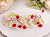 The "Pack of 5 mini scrunchies with cherry hanging" by Pret my baby features playful red pom-poms, pastel animal beads, white lace, and ruffled elastic—perfect for brightening up any hairstyle or braid. Displayed on a white plate with flowers in the background.