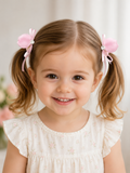 A young girl with light brown hair smiles warmly indoors, wearing Pret my baby's satin tulip bud fascinator hairclips with ribbon trails and a white, ruffled, floral dress against a soft, neutral background.