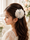 A young girl with long wavy brown hair, wearing a white lace dress and the Pret my baby Rhynestone butterfly on lace bow fascinator hairclip, looks to the side in a softly lit, floral setting.