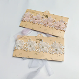 The Pret my baby Lace headband features soft flowers and pearl accents on a lace band, displayed on cream cards with gold designs. Available with light pink or white flowers and tulle ribbons—an elegant accessory for any outfit.
