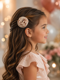A young girl with long, wavy brown hair wears a shiny pink dress and a Pret my baby Satin rose fascinator clip adorned with pearls, smiling in profile against a softly blurred background with pink and white balloons.