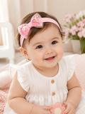 A happy baby wearing a white dress with wooden buttons and the “Dancing confetti bow soft baby headband” by Pret my baby sits indoors, surrounded by a pink blanket and flowers in the background.