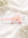 The Fancy Fascinator Little Tulips Baby Headband by Pret my baby, featuring a pink tulle bow with flower accents, rests on a Fashion Jewelry card amid baby's breath on soft cream fabric.