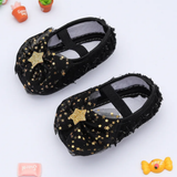 The Pret my baby Rosette bellies with star - black are baby girl shoes adorned with sparkly gold stars and a gold star embellishment, featuring black elastic straps and mesh fabric, shown surrounded by decor on a white background.
