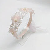 The Pret my baby Lace headband with soft flowers and pearls features a delicate lace band adorned with light pink flowers and small pearl accents, shown on a clear stand against a plain white background.