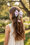 A young girl with long, wavy brown hair wears a Pret my baby Medium fascinator tulip bow in her hair and a white sleeveless dress, standing in a sunlit garden filled with blurred tulips and lush greenery.