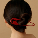 A person with dark brown hair in a low bun, secured with the Pretmebabe Safety Pin bun clip—a large red metal hair fork—against a plain beige background.