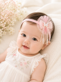 A happy baby in white lies on a cream blanket beside delicate white flowers, wearing the Pret my baby Fancy fascinator crystal butterfly on bow headband—perfect for highlighting adorable infant hair accessories.