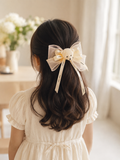 A young girl with long, wavy dark hair wears the Pret my baby Statement bow with teddy bear—a pastel tulle hair clip with a cute bear face and crystal center—paired with a cream dress, standing indoors by a vase of flowers.