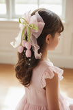 A young girl with wavy brown hair wears a pink dress and the Pret my baby Tulip with pearls large bow fascinator hairclip, featuring satin tulip buds, green ribbon, and pearls, while standing in a bright room.