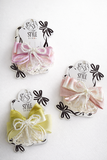 Three Multiple bow with doiley lace fascinator hairclips by Pret my baby in pink, purple, and yellow are displayed on branded cards, each featuring pearls and attached to alligator pins on a white textured surface.
