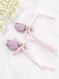 The Pret my baby Pair of satin tulip bud fascinator hairclips with ribbon trails features purple buds, pink bows, and gems, styled with a baby pink lace bow. Shown on a white background beside delicate white baby's breath flowers.