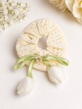 The Tulip bow lace scrunchie by pretmebabe features cream cotton lace, a green ribbon, a white flower charm, and two white tulip bows, displayed on a white surface with delicate flowers in the background.