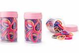 Three clear plastic jars with pink lids, labeled "Pret my baby Pack of 100 everyday mix hair ties," feature colorful soft hair ties inside. Two jars stand upright and one lies on its side, spilling hair ties onto a white surface.