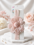The Fascinator extra large flower with rose bud baby headband by Pret my baby is shown on a white card atop marble, with soft-focus pink and white flowers in the background.