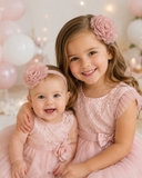 Two young girls in matching mauve dresses and pretmybaby’s Flower Bloom Fascinator Sibling Set smile together. The older hugs the younger, who sits happily among softly lit pink and white balloons.