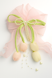 The Tulip with Pearls Large Bow Fascinator Hairclip by Pret my baby features a pink sheer ribbon bow adorned with three pastel satin tulip buds, green stems, pearl strands, and heart accents—a delicate handcrafted hair accessory.