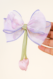 A hand holds the Pret my baby Medium fascinator tulip bow—a decorative organza accessory in sheer light pink with purple edges, featuring a green ribbon and a pink rosebud ornament, ideal for pairing with fresh tulips.