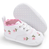 A pair of Pret my baby Embroidery sneakers - white, featuring pink and white flower embroidery, pink scalloped trim, and white laces. One pre-walker stands upright while the other shows its polka-dotted sole.