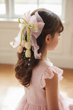 A young girl with wavy brown hair wears a light pink dress and the Pret my baby Tulip with pearls large bow fascinator hairclip, featuring satin tulip buds, pastel ornaments, and pearls, facing away in a softly lit room.